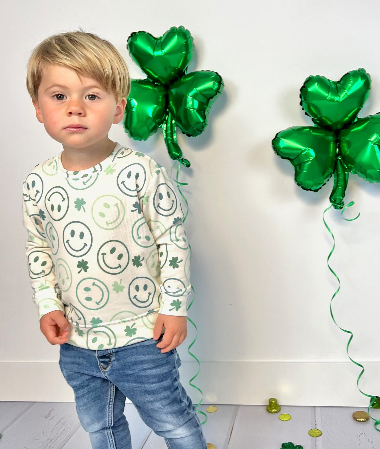 A child wearing a long-sleeved white sweatshirt with a pattern of green smiley faces and clovers, standing next to green clover-shaped balloons.
